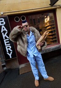 Ember hosszú műszőrme kabátban, világoskék pizsamában, barna papucsban, kezében papírba csomagolt bagettekkel, egy pékség előtt áll.