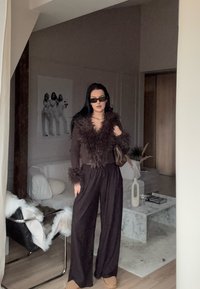 Top maro cu dungi și guler și manșete pufoase, asortat cu pantaloni negri largi. Ochelari de soare negri și o geantă maro, cu un fundal interior minimalist.