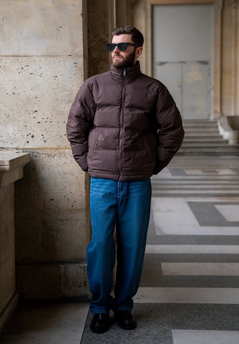 Homme barbu portant des lunettes de soleil, une doudoune sombre, un jean bleu et des chaussures noires, debout les mains dans les poches dans un corridor en pierre.