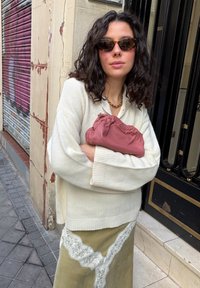 Femme aux cheveux bouclés portant des lunettes de soleil, un pull crème, une jupe en dentelle olive, tenant une pochette rose plissée, debout sur un trottoir en ville.