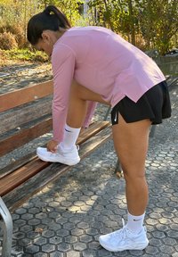 Scarpe sportive bianche con rete testurizzata, una maglietta a maniche lunghe leggera di colore rosa e pantaloni corti neri plissettati, indossati mentre si è seduti su una panchina di legno.
