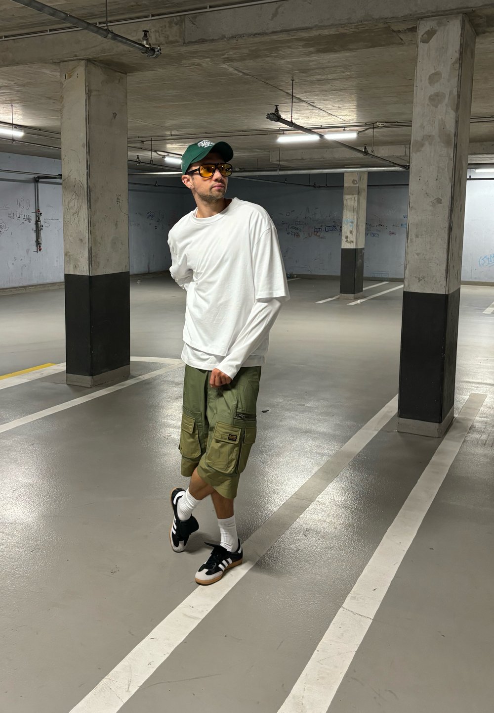 Witte longsleeve shirt over een short-sleeve shirt, groene cargoshorts, zwarte sneakers met witte accenten en gele getinte zonnebril.