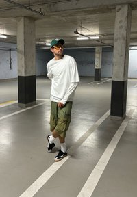 Witte longsleeve shirt over een short-sleeve shirt, groene cargoshorts, zwarte sneakers met witte accenten en gele getinte zonnebril.