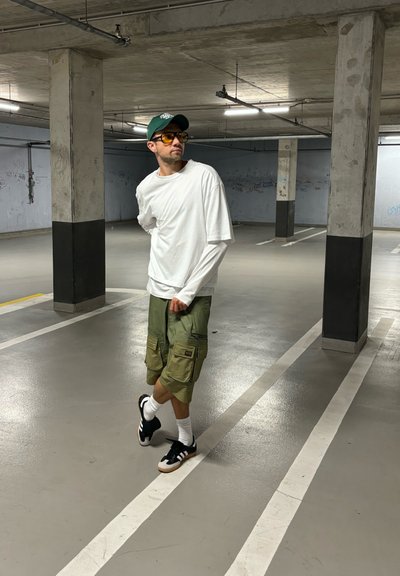 Camisa blanca de manga larga superpuesta a una camisa de manga corta, pantalones cortos cargo verdes, zapatillas negras con acentos blancos y gafas de sol con tintado amarillo.