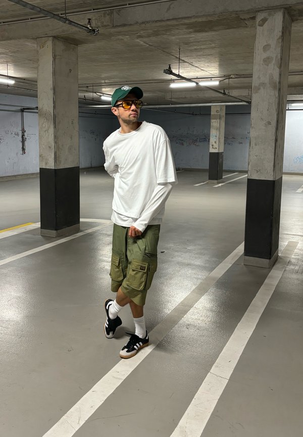White long-sleeve shirt layered over a short-sleeve shirt, green cargo shorts, black sneakers with white accents, and yellow tinted sunglasses.
