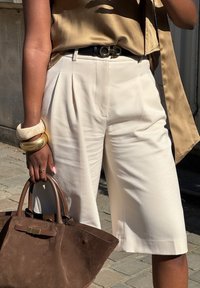 Pantalones cortos de talle alto plisados en color beige claro con un corte relajado, combinados con una blusa de satén dorada y un bolso de ante marrón. Los accesorios incluyen pulseras apiladas.