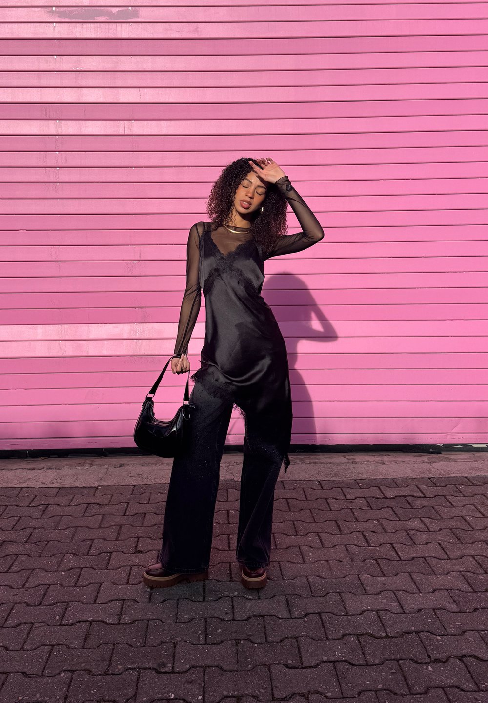 Femme portant un haut noir à manches en voile et un pantalon noir, tenant un sac à main, debout sur des pavés devant un mur rose vif, se protégeant les yeux du soleil.