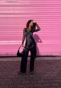 Femme portant un haut noir à manches en voile et un pantalon noir, tenant un sac à main, debout sur des pavés devant un mur rose vif, se protégeant les yeux du soleil.