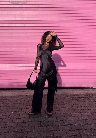 Mujer con blusa negra de mangas transparentes y pantalones negros sostiene un bolso, de pie sobre adoquines frente a una pared de color rosa brillante, protegiéndose los ojos del sol.