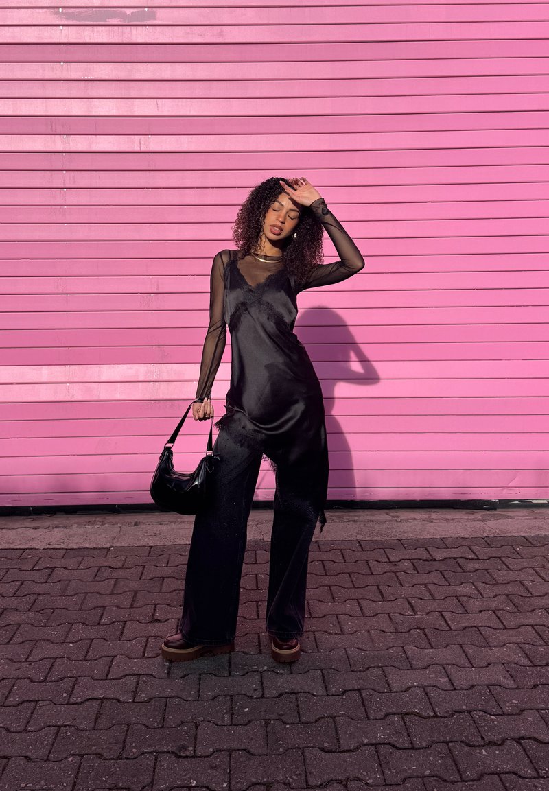 Frau in schwarzem, durchsichtigärmeligem Oberteil und schwarzer Hose hält Handtasche, steht auf Kopfsteinpflaster vor einer knallpinken Wand und schützt die Augen vor der Sonne.