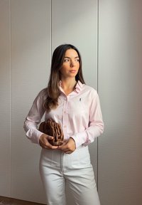 Světle růžová košile na knoflíky s dlouhými rukávy a vyšívaným logem, v kombinaci s bílými kalhotami s vysokým pasem a hnědou tkanou psaníčkem.
