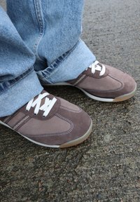 Baskets en daim marron avec des lacets blancs portées avec un jean en denim bleu clair, debout sur un sol en asphalt rugueux.