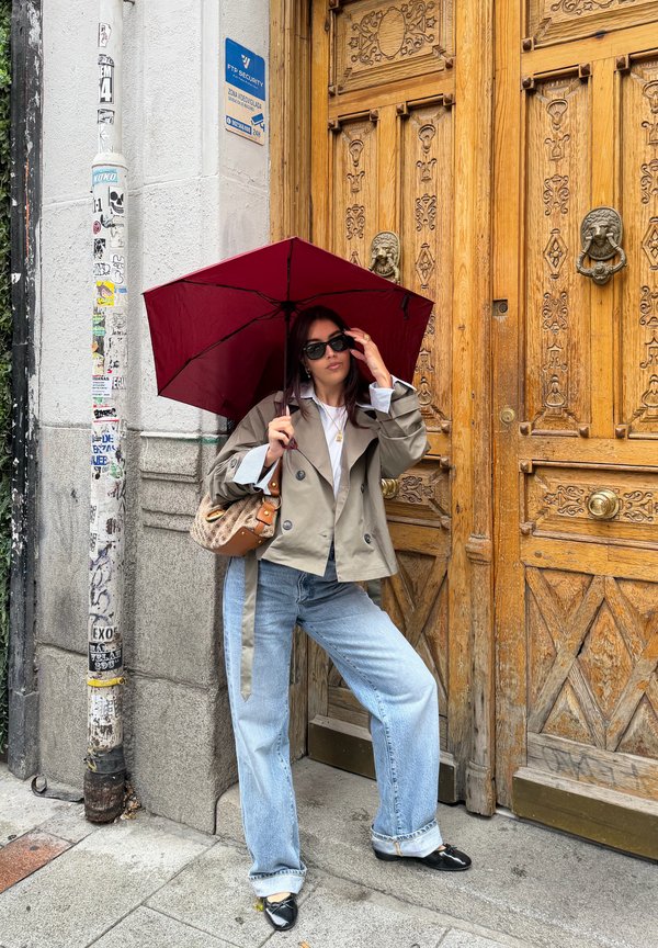 Una mujer con gafas de sol sostiene un paraguas color burdeos delante de una puerta de madera ornamentada en una calle de la ciudad.