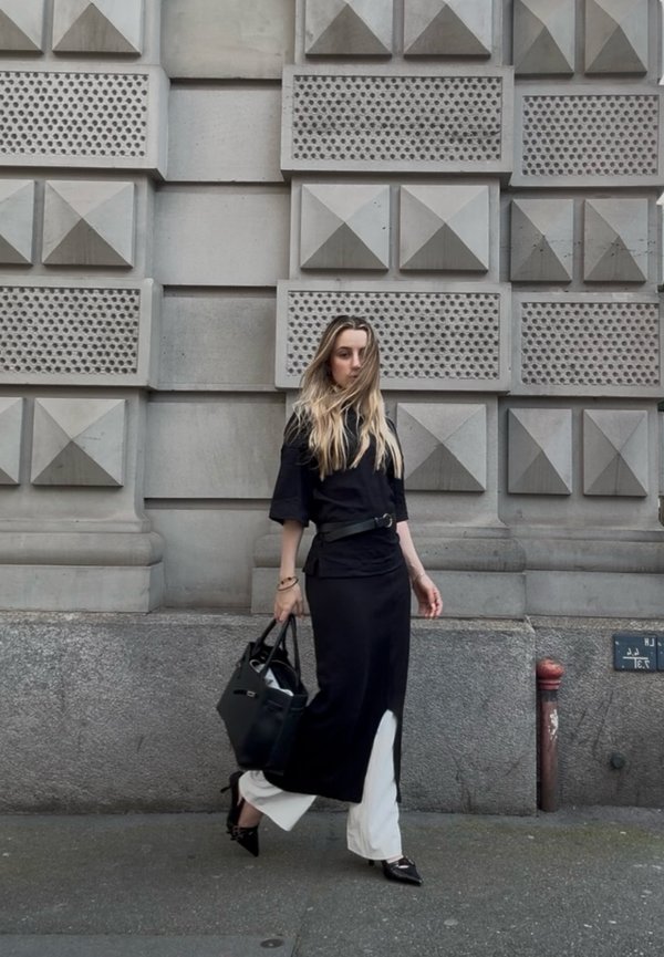 Woman with long blonde hair wearing black top and skirt over white pants walks past textured stone wall holding black handbag.