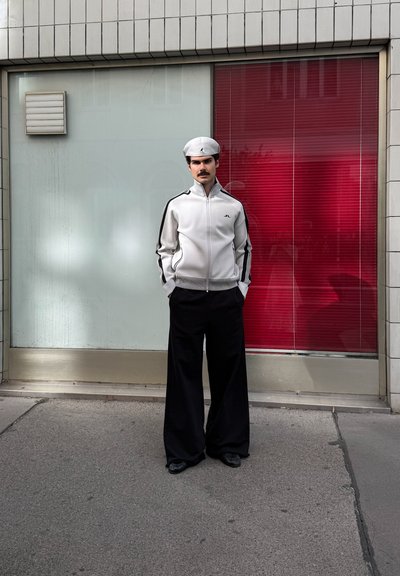 Chaqueta deportiva en gris claro con rayas negras, combinada con pantalones amplios negros. Una gorra blanca completa el conjunto. El fondo presenta una ventana roja.