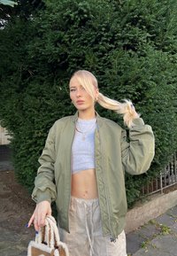 Chaqueta bomber verde, top corto gris, pantalones baggy beige y un bolso mullido beige. Pelo largo rubio recogido en una coleta. Fondo de follaje verde.