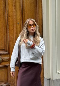 Szary sweatshirt, brązowa spódnica i okulary przeciwsłoneczne w odcieniu pomarańczowym. Trzyma brązową torbę z dużym, błyszczącym dodatkiem do włosów na nadgarstku. Drewniane tło.
