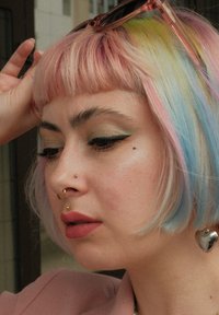 Kort rosa hår med blå og grønne høydepunkter, gullpiercinger i nesen og hjerteformet ørering, iført en rosa blazer og solbriller.
