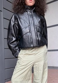 Fekete, rövidített bőrkabát magas gallérral és első zsebekkel. Texturált felület és kontrasztos cipzár részlet. Világos bézs nadrággal párosítva.