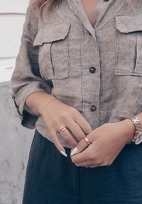 Koszula na krótki rękaw, zapinana na guziki, wykonana z teksturowanego jasnoszarego lnu, z dwiema kieszeniami na piersi. Ręce ozdobione wieloma pierścionkami, noszące zegarek.