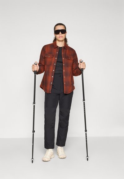 Camisa de franela a cuadros rojos y negros sobre ropa de deporte negra. Bastones de trekking negros, zapatos ligeros y pantalones oscuros con parches en las rodillas.