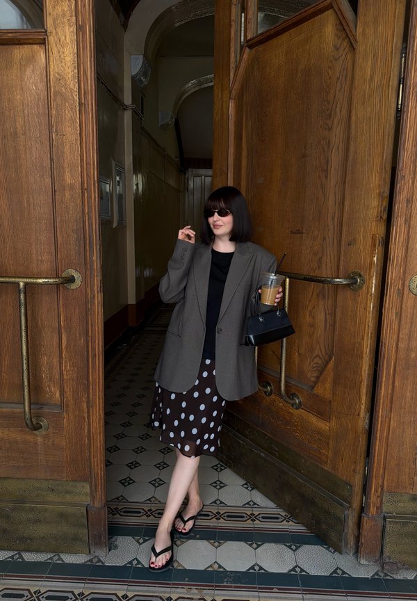 Femme portant des lunettes de soleil, un blazer oversize et une jupe à pois, debout dans un encadrement de porte en bois ouvert, tenant un café glacé et un sac à main noir.