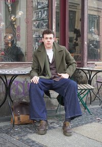 Casaco verde de lã por cima de um colete às quadrículas, t-shirt branca e jeans azuis largos. Botas de couro castanhas e uma mala de carteiro castanha ao lado.