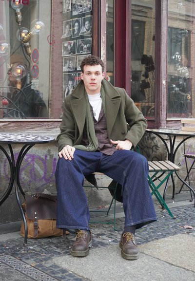 Abrigo verde de lana sobre un chaleco a cuadros, camiseta blanca y vaqueros azules holgados. Botas de cuero marrón y una bolsa marrón de mensajero al lado.