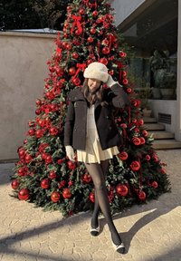 Cappotto di lana nero e cappello di pelliccia bianco, gonna plissettata gialla, collant neri e scarpe bicolore, posata davanti a un albero di Natale decorato.