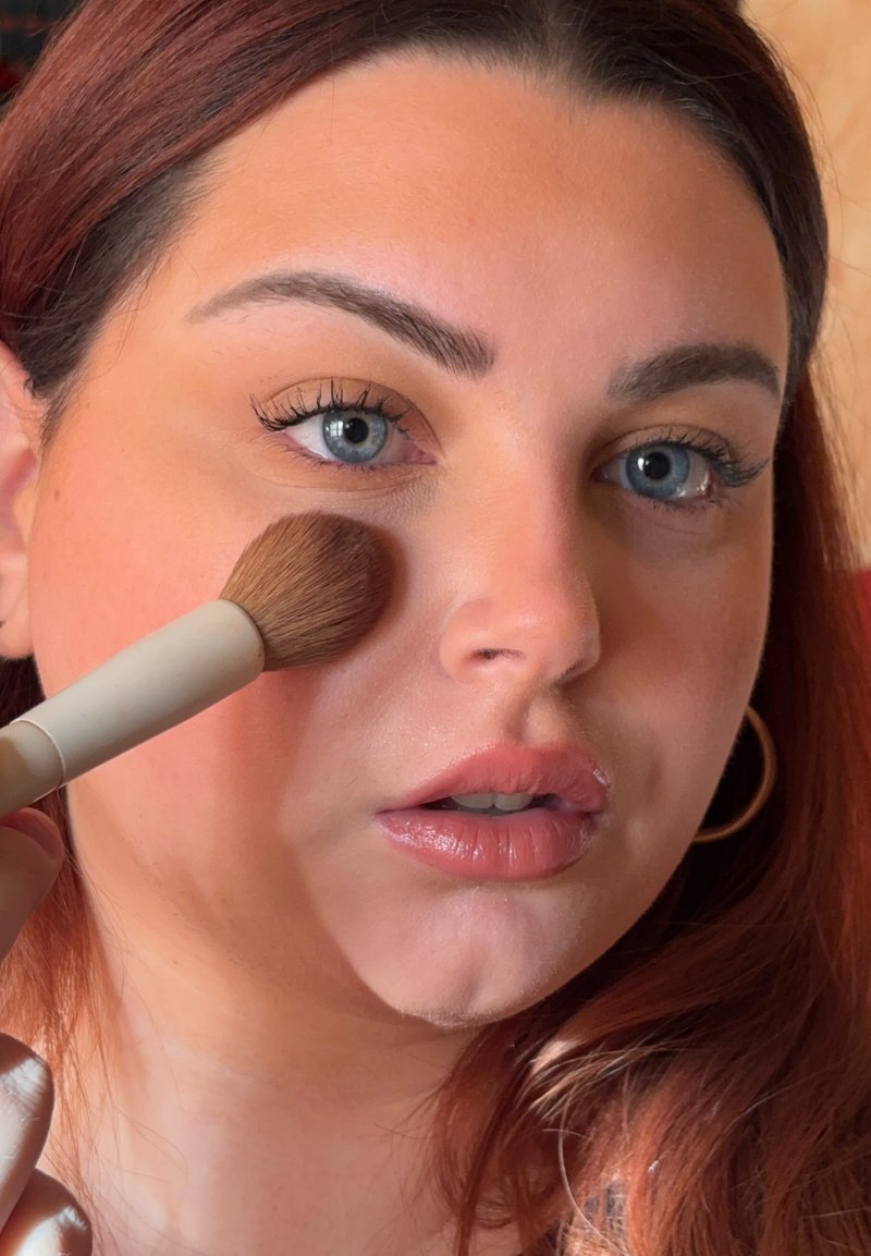 Model brengt blush aan met een ronde make-upkwast. Heeft een zachte huid, volle lippen en lange wimpers. Het haar is golvend en kastanjebruin.