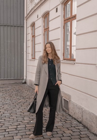 Abrigo de lana gris claro sobre una blusa oscura, combinado con pantalones negros y zapatos grises. El bolso es de cuero negro, con una textura suave.