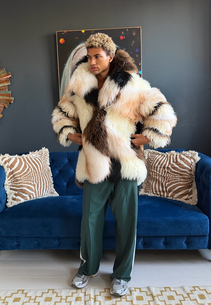 Personne aux cheveux blonds bouclés portant un manteau en fourrure duveteuse multicolore, un pantalon vert et des baskets, debout dans un salon moderne.