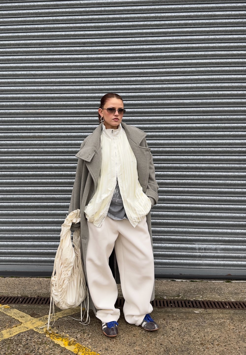 Long manteau gris, haut plissé blanc, chemise grise, pantalons blancs surdimensionnés, chaussures bleues, sac surdimensionné, sur fond de volet roulant métallique.