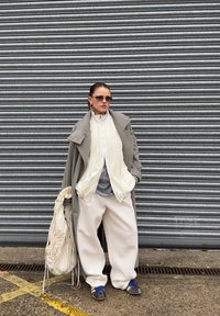 Long manteau gris, haut plissé blanc, chemise grise, pantalons blancs surdimensionnés, chaussures bleues, sac surdimensionné, sur fond de volet roulant métallique.