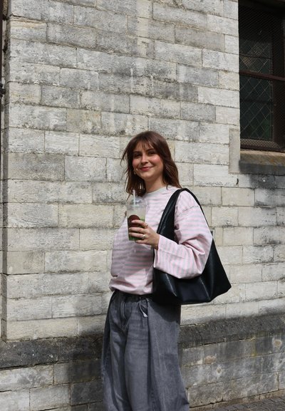 Joven con cabello castaño que lleva un suéter rosa a rayas y pantalones grises, sosteniendo una bebida verde y un bolso negro de hombro junto a un muro de piedra.
