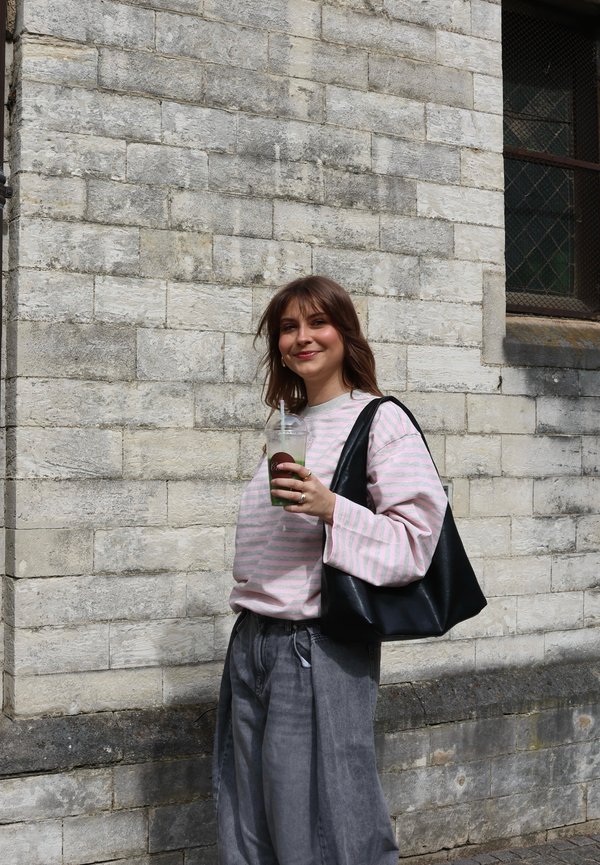 Ung kvinde med brunt hår iført en pink stribet sweater og grå bukser, der holder en grøn drink og en sort skuldertaske ved en stenmur.