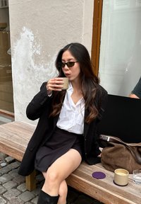 Blazer nero sopra una camicia bianca con bottoni e una gonna nera. Tiene in mano una tazza marrone. Panchina di legno, borsa scura e sottobicchieri circolari visibili.