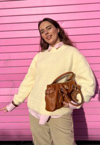 Pull en tricot crème avec épaules tombantes, porté sur une chemise à col rose clair. Sac à main en cuir marron avec détails à boucle.