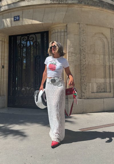 Falda maxi texturizada blanca, camiseta gráfica con diseño de fresa, zapatillas rojas, sosteniendo un casco blanco y un bolso rojo. Gafas de sol negras.