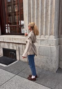 Lichtbeige jasje met riem en lange mouwen, gecombineerd met een blauwe jeans en bruine schoenen. Een vrouw staat bij een stenen muur met een drankje in haar hand.