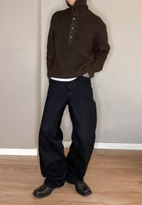 Pull-over en maille côtelée marron avec trois boutons noirs, associé à un pantalon large noir et des bottines noires, sur un sol en bois.