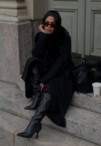 Fekete tűsarkú combcsizmák, hosszú fekete kabátot és szőrme gallért viselve, kőlépcsőn ülve, mellette egy fekete kézitáska.