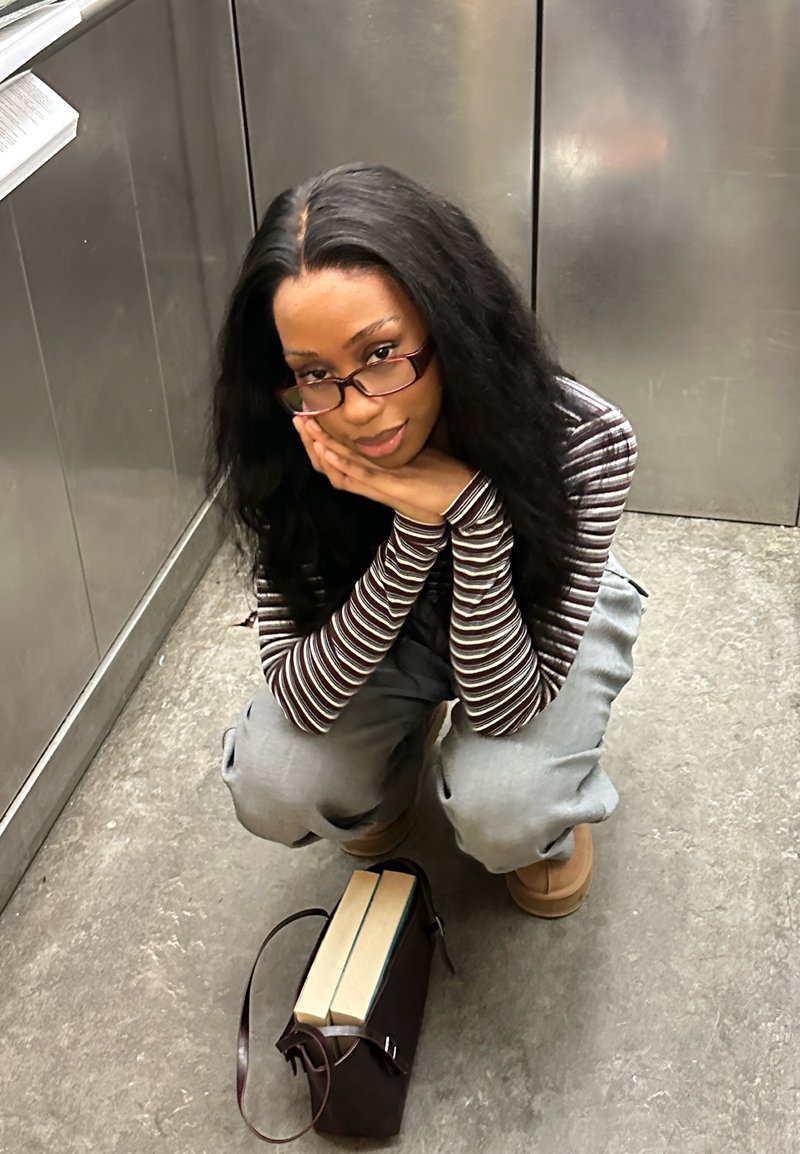 Frau mit langen Haaren und Brille hockt auf einem Betonboden, stützt ihr Gesicht auf die Hände, vor ihr eine braune Tasche und zwei Bücher.