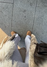 Bottes en daim marron, pantalon de jogging gris et un manteau avec des accents de fourrure, debout sur un pavé texturé gris sous une lumière douce.