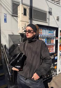 Casaco bomber preto com colarinho alto, óculos de sol e uma bolsa preta texturizada. Ambientado diante de uma máquina de venda automática.