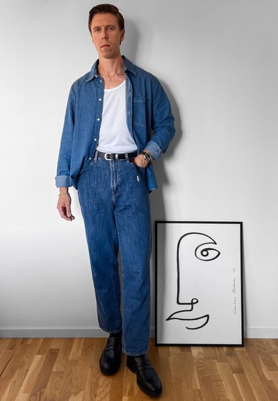 Camisa de mezclilla y jeans en azul, camiseta blanca de tirantes, cinturón negro, zapatos de cuero negros. Obra de arte abstracto de cara enmarcada en la pared. Suelo de madera.