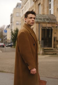 Long manteau en laine marron avec un design croisé, de grands revers et des poignets boutonnés. Accessoires comprennent un sac beige et une bague.