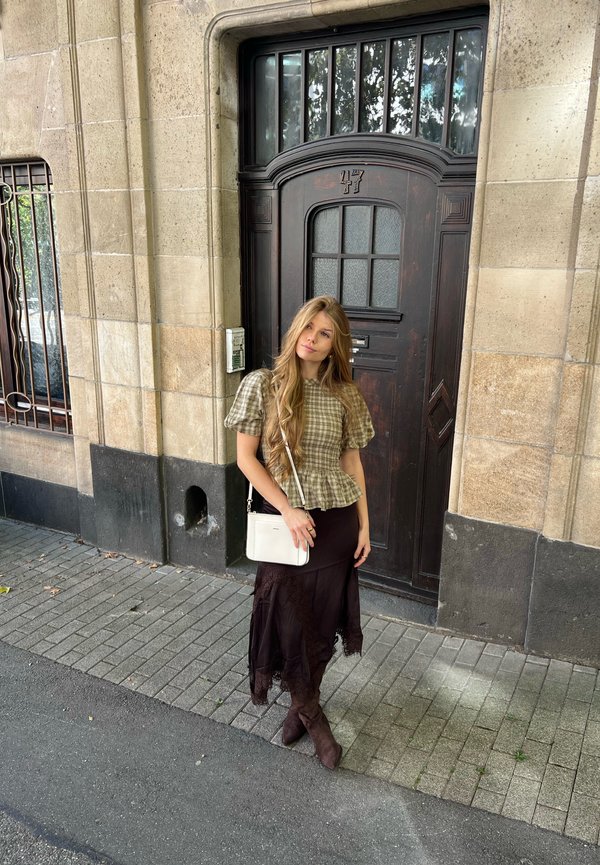 Jeune femme aux longs cheveux portant un chemisier à carreaux et une jupe foncée, debout sur le trottoir devant une porte en bois foncé numérotée 47 dans un bâtiment en pierre.