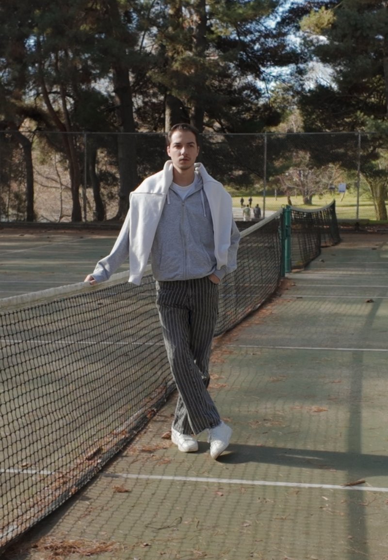 Junger Mann in grauem Hoodie und gestreifter Hose lehnt an einem Tennisnetz auf einem Außenplatz, umgeben von Bäumen an einem sonnigen Tag.
