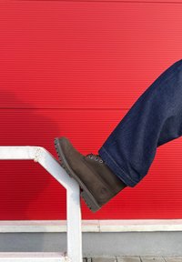 Botte en cuir marron à bout rond, laçage avec œillets et semelle texturée, posée sur une rambarde blanche. En arrière-plan, un panneau rouge.